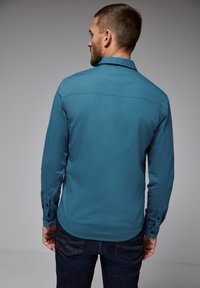 Tyrkysová košile s dlouhým rukávem a knoflíkovými manžetami, klasickým límcem a zaobleným lemem, nošená s tmavými džínami. Hladká textura, bez vzorů.