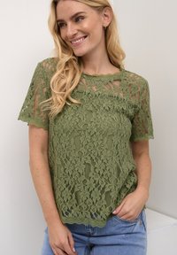 Top in pizzo verde oliva con maniche corte, scollatura ondulata, motivi floreali e una vestibilità ampia, abbinato a jeans di denim azzurri chiari.