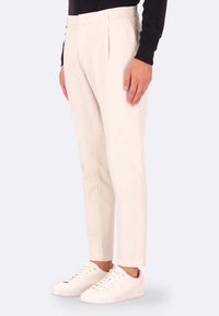 Pantaloni tapered beige con una parte anteriore plissettata, realizzati in tessuto misto cotone, abbinati a sneaker bianche basse per un look casual.