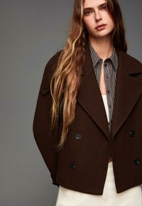 Veste double boutonnage en mélange de laine marron avec de grands boutons noirs, revers larges et coupe décontractée, portée sur une chemise rayée.