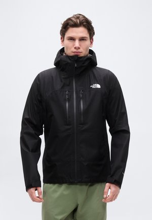 Mladý muž v čiernej kapucňovej bunde North Face s dvoma zipsovými vreckami na hrudi a zelených nohaviciach, stojaci pred čistým bielym pozadím.