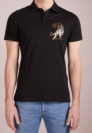 Homme portant un polo noir avec un graphisme de tigre doré et le texte « Versace Jeans », assorti à un jean bleu, debout les mains détendues.