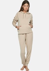 Vrouw met lang bruin haar draagt een beige hoodie en bijpassende joggingsbroek, staat blootsvoets met haar handen in de zakken van de hoodie.