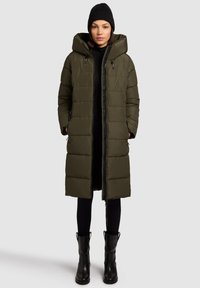 Olivenfarvet lang polstret frakke med hætte, der har en lynlås foran, sidelommer og en glat tekstur. Bæres med sorte leggings og støvler.