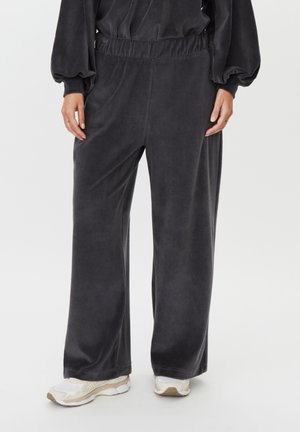 Persona con pantalones de terciopelo negro holgados y zapatillas blancas, de pie frente a un fondo claro y liso.
