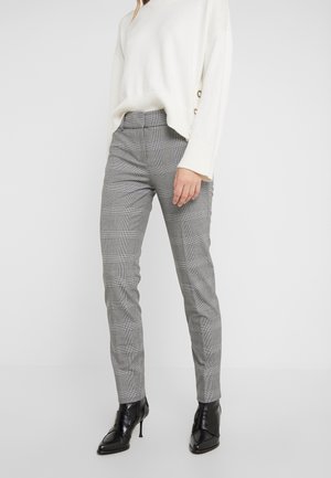 Frau trägt graue karierte Slim-Fit-Hosen, weißen Langarmpullover mit seitlichen Knopfdetails und schwarze spitze, hochhackige Stiefeletten.
