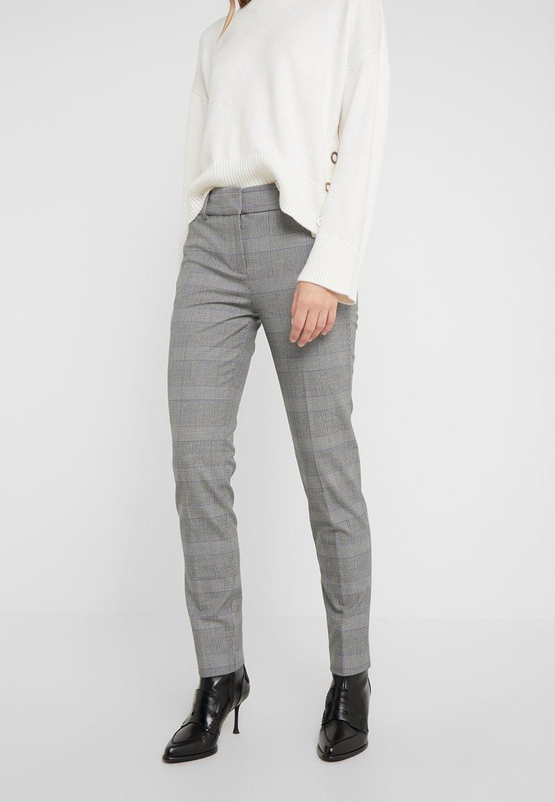 Femme portant un pantalon slim à carreaux gris, un pull blanc à manches longues avec des boutons sur le côté, et des bottines noires à talons hauts pointues.