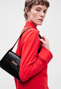 Μαύρη δερμάτινη τσάντα ώμου με κούμπωμα καπάκι, χρυσές λεπτομέρειες και μακριά λουρί. Το μοντέλο φοράει ένα ζωντανό κόκκινο μπλουζάκι.