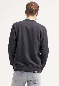Homme de dos portant un sweat-shirt gris foncé et un jean bleu clair sur un fond blanc uni.