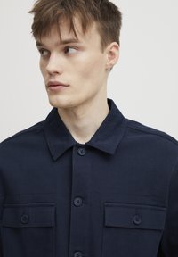 Veste boutonnière bleu marine en tissu texturé. Elle possède un col et deux poches poitrine avec des fermetures à bouton.