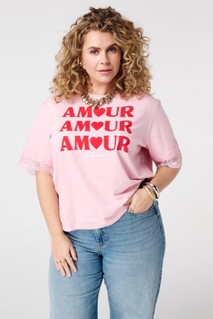 Gekrulde blonde vrouw die een roze T-shirt draagt met rode "AMOUR" tekst en hartontwerpen, blauwe spijkerbroek, gouden ketting en armbanden, poserend tegen een witte achtergrond.