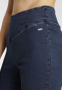 Dunkelblaue hochgeschnittene Jeans mit glatter Oberfläche, dezenten Nähten und einem kleinen Metalllogo-Akzent auf der Vorderseite.