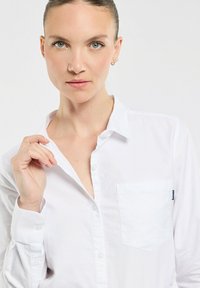 Chemise blanche à manches longues en coton avec une poche sur le côté gauche. Le col est légèrement ouvert, montrant une texture lisse et un design épuré.