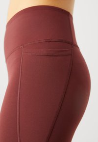 Burgunderfargede leggings laget av elastisk stoff, med høy midje, flate sømmer og en liten baklomme for praktisk bruk.