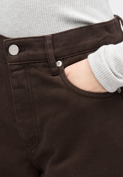 A barna farmernadrág magasított derékrésszel, ötlovas zsebes kialakítással és ezüstszínű fémrészletekkel rendelkezik, texturált anyagból készült, és látható varrásokkal.