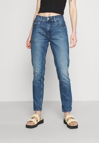 Jeans de tiro alto azul desgastados con un diseño de cinco bolsillos. Combinados con sandalias amarillas claras con volantes sobre una suela negra plana.