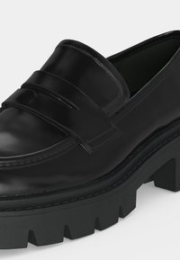 Μαύρο δερμάτινο loafer με χοντρή εξωτερική σόλα και κλασική λεπτομέρεια λουριού στο πάνω μέρος, εμφανίζεται από γωνία κοντινής λήψης.