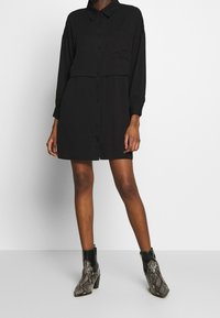 Vestito camicia nero a maniche lunghe con chiusura a bottoni e taschino sul petto; abbinato a stivali alla caviglia con motivo a serpente grigio e nero.