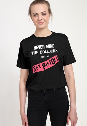 Młoda kobieta w czarnej koszulce z białym i różowym napisem "Never mind the bollocks, here's the Sex Pistols,", stoi na jednolitym tle.