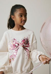 Lys beige sweatshirt med et stort lyserødt og gyldent pailletbånddesign. Ærmerne er voluminøse, og stoffet ser blødt ud.