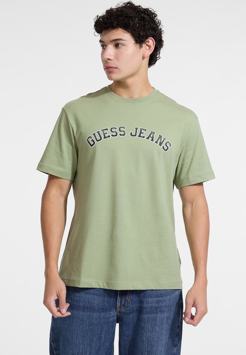 Alyvuogių žalia medvilninė marškinėliai su užrašu "GUESS JEANS" nespalvotai spausdintu per krūtinę. Trumpomis rankovėmis ir apvaliu iškirpimu. Kasdienis siluetas.