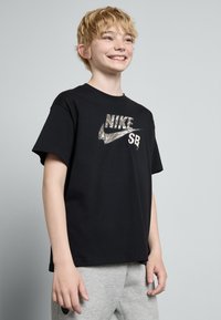 Černé bavlněné tričko s metalickým stříbrným logem Nike a růžovým textem "SB", s volným střihem a krátkými rukávy.