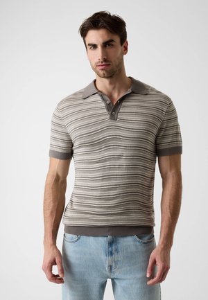 Homme debout devant un fond uni, portant un polo en maille gris à rayures et des jeans bleu clair, face à l'avant.