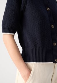 Cardigan texturé bleu marine à manches courtes avec boutons, ourlet côtelé et bordure blanche, porté avec un pantalon beige clair, main dans la poche.