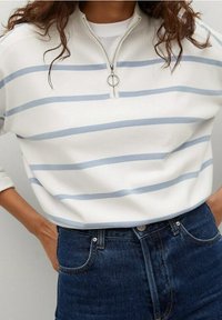 Felpa bianca con zip e righe orizzontali blu, dotata di un colletto semi-alto e in tessuto morbido, abbinata a jeans di denim scuro.