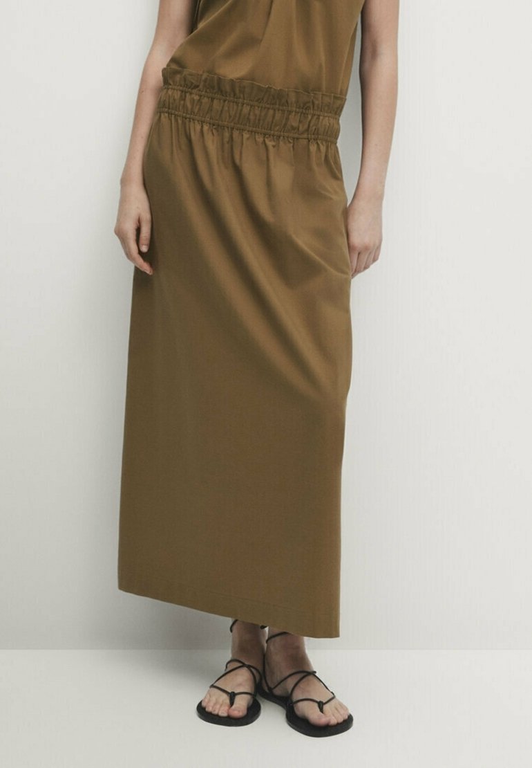 Femme porte une jupe midi marron avec une taille élastique et des sandales plates noires à brides, debout devant un fond clair uni.
