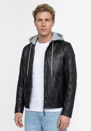 Jonge man met krullend blond haar, gekleed in een zwarte leren jas met een grijze capuchon, wit t-shirt en lichtblauwe spijkerbroek, kijkt recht vooruit.