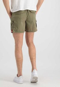 Cargo-Shorts aus olivgrünem Stoff, mit zwei Seitentaschen, normaler Passform und einem kleinen roten Logo-Aufnäher. Tragen Sie sie mit weißen Turnschuhen.