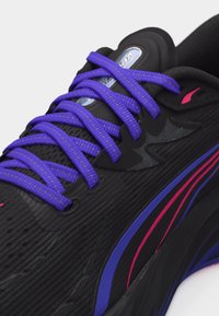 Chaussure de sport noire avec des lacets violets, des rayures roses et violettes sur le côté, et un logo "Vera" visible sur la languette.