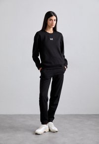 Felpa nera e pantaloni sportivi realizzati in tessuto morbido con una vestibilità comoda. Presentano un piccolo logo sul petto e polsi a costine. Indossati con sneakers bianche.