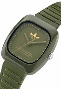 Montre en silicone verte avec un cadran carré, des accents dorés et un logo à trois rayures. Elle possède un bracelet texturé et des aiguilles des heures et des minutes simples.