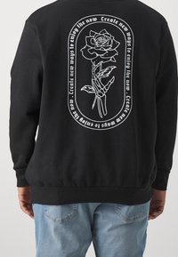 Sudadera negra con un gráfico blanco de una mano esquelética sosteniendo una rosa y el texto "Crea nuevas formas de disfrutar el ahora" en un marco circular.