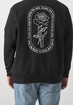 Sweatshirt noir avec un graphique blanc représentant une main squelettique tenant une rose et le texte "Créez de nouvelles façons de profiter de l'instant présent" dans un cadre circulaire.