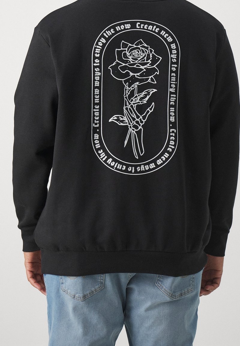 Sudadera negra con un gráfico blanco de una mano esquelética sosteniendo una rosa y el texto "Crea nuevas formas de disfrutar el ahora" en un marco circular.