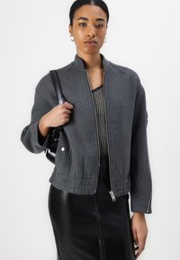 Grau zip-up Jacke aus strukturiertem Stoff mit elastischem Saum, seitlichen Taschen und abgesenkten Schultern. Über einem gestreiften Oberteil getragen und mit einem schwarzen Rock kombiniert.
