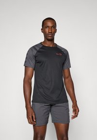 T-shirt de sport noir avec des manches raglan grises, arborant un logo rouge sur la poitrine, associé à un short gris, en tissu lisse et à coupe active.