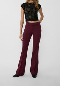 Czarny koronkowy crop top z krótkimi rękawami, zestawiony z dopasowanymi bordowymi spodniami w fasonie dzwonów. Czarne buty dopełniają outfit. Widoczne proste akcenty biżuteryjne.