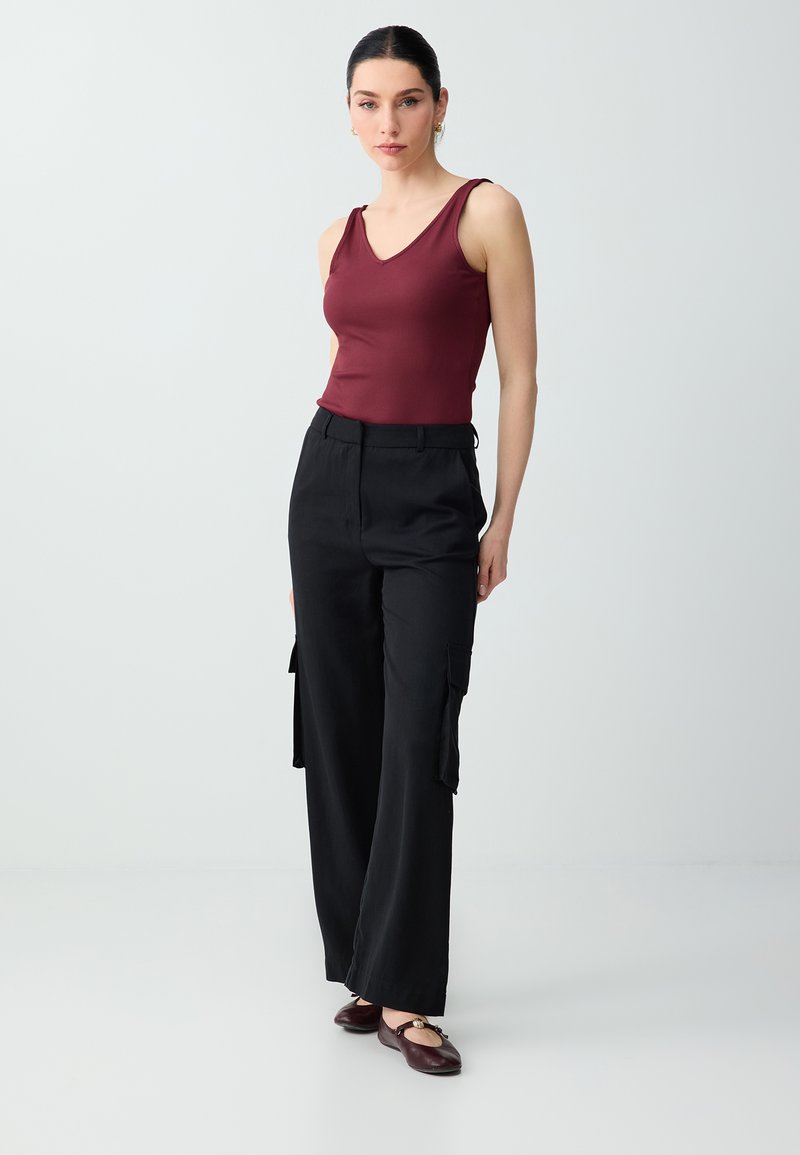 Femme portant un haut bordeaux sans manches, un pantalon cargo noir à jambes larges et des ballerines bordeaux, debout devant un fond clair uni.