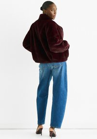 Woman in burgundy faux fur jacket, blue high-waist jeans, and dark brown heels standing against a plain white background, looking over shoulder.