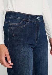 Gros plan sur un jean en denim bleu foncé avec des coutures orange, fermeture à bouton, poches avant, porté avec un haut à manches longues blanc.