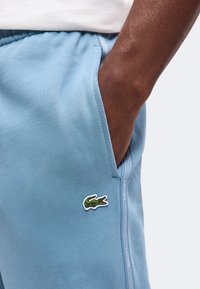 Χέρι στην τσέπη του ανοιχτό μπλε φόρμας Lacoste με το λογότυπο κροκόδειλου στο μηρό και άσπρη πλαϊνή ρίγα με το όνομα της μάρκας.