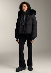 Čierna vatovaná bunda s kapucňou lemovanou kožušinou, so zúženým strihom, nadmerným rukávom a pod ňou priliehavý sveter s vysokým golierom na zips.