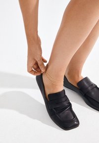 Woman wearing black leather loafers adjusts delicate double gold anklet on bare ankle against white background.
