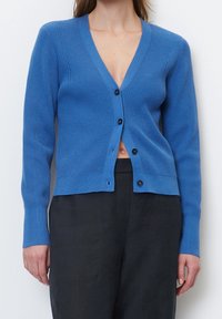 Žena v modré žebrované kardiganu na knoflíky a černých kalhotách s vysokým pasem, ukazující malou mezeru na břiše pod knoflíky.