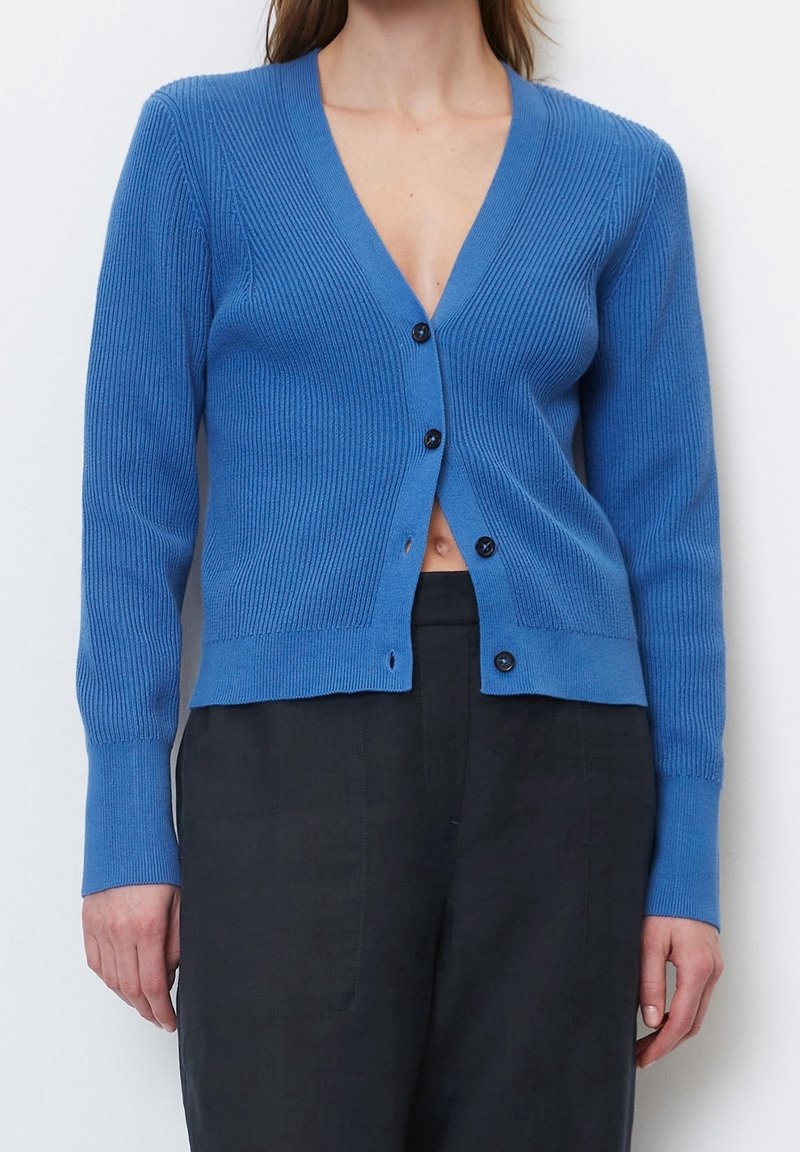 Žena v modré žebrované kardiganu na knoflíky a černých kalhotách s vysokým pasem, ukazující malou mezeru na břiše pod knoflíky.