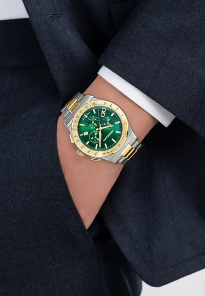 Randmepael roheka rohelise sihverplaadi, kuldsete aktsentide ja hõbe-kulla metallrihmaga, kantud vasakul randmel tumelanahalise ülikonnajaki ja valge särgi varrukamanseti all.